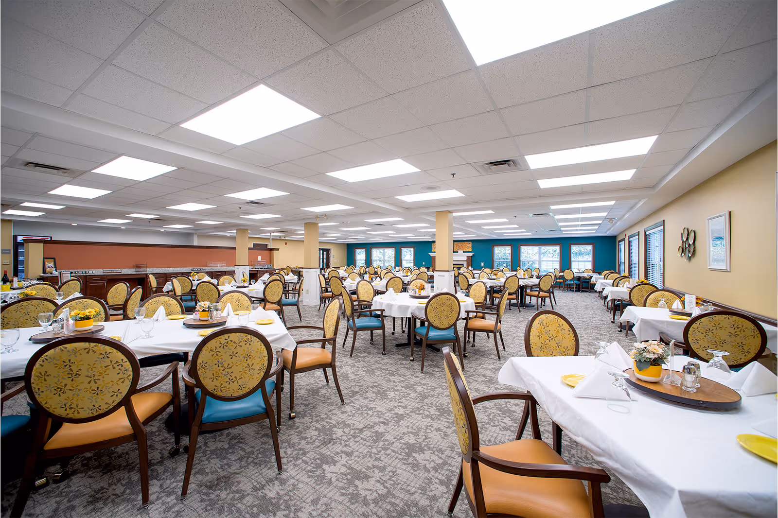 A spacious dining room with multiple round and rectangular tables covered with white tablecloths. Each table is set with folded white napkins, glassware, and small flower arrangements. The chairs have wooden frames with patterned yellow upholstery. The room has a carpeted floor, beige and teal walls, and large windows letting in natural light.