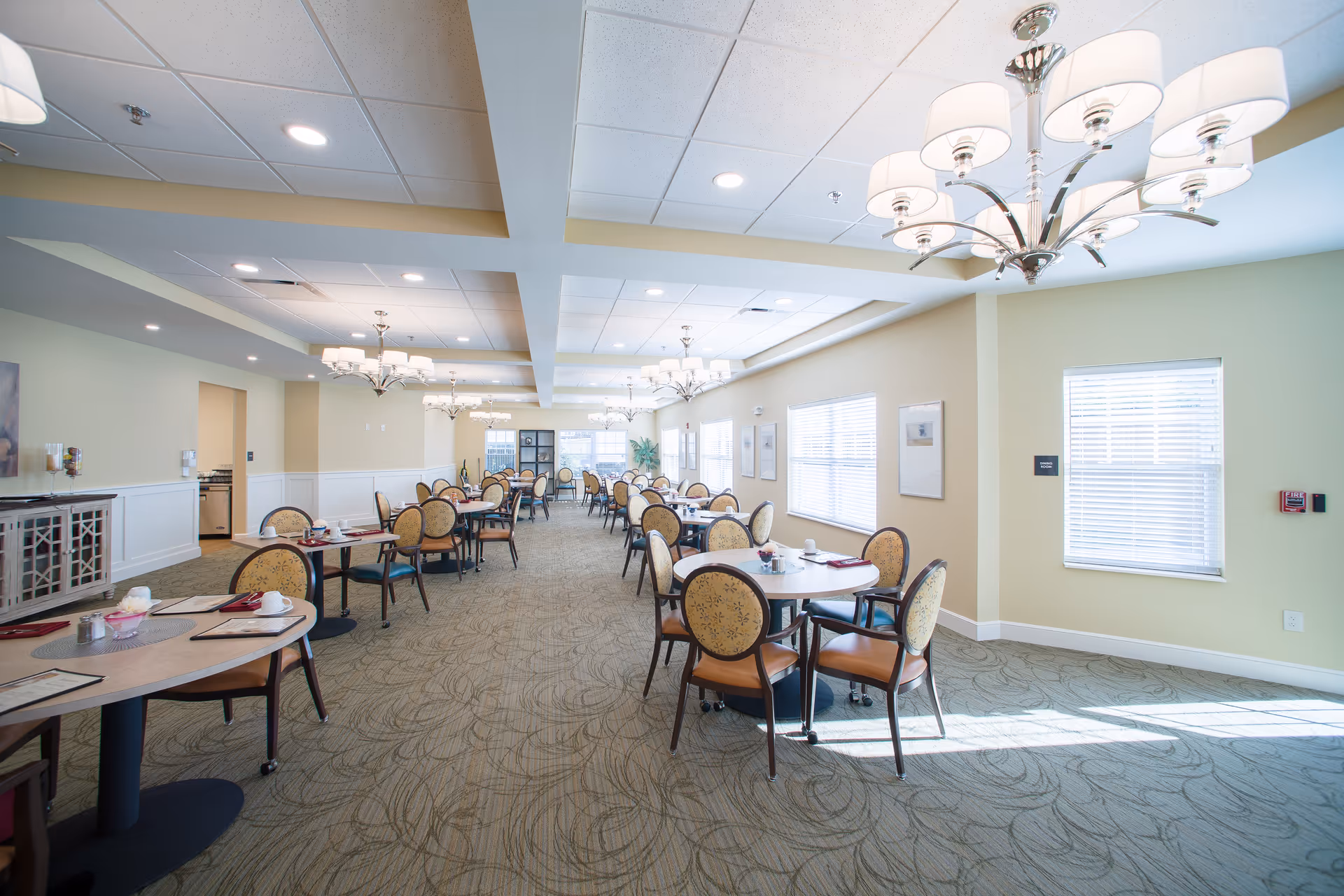 Bright and spacious dining room with multiple round tables and chairs arranged neatly. The room features large windows with blinds allowing natural light to fill the space, elegant chandeliers hanging from the ceiling, and light-colored walls with framed artwork. The carpet has a subtle pattern, and there is a sideboard cabinet along one wall.