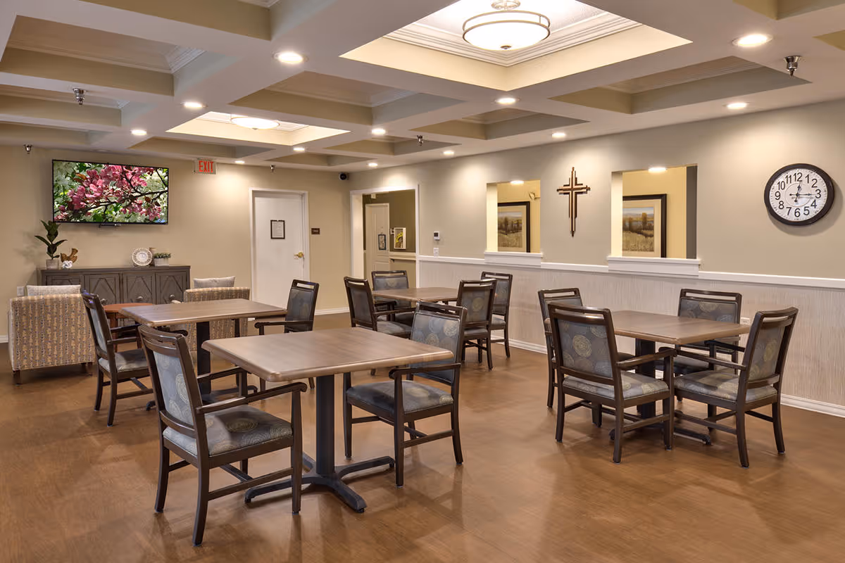 Well-lit communal dining room with multiple wooden tables and chairs, a wall-mounted TV, clock, and decorative furnishings.