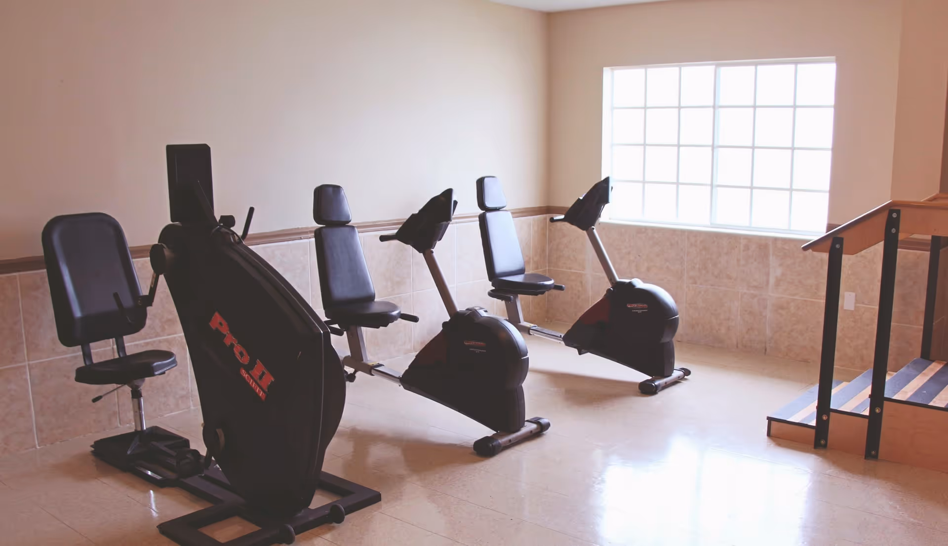 A small exercise room with three stationary recumbent bikes positioned near a large window that lets in natural light. The room has tiled walls and a beige floor, with a wooden handrail and steps on the right side.
