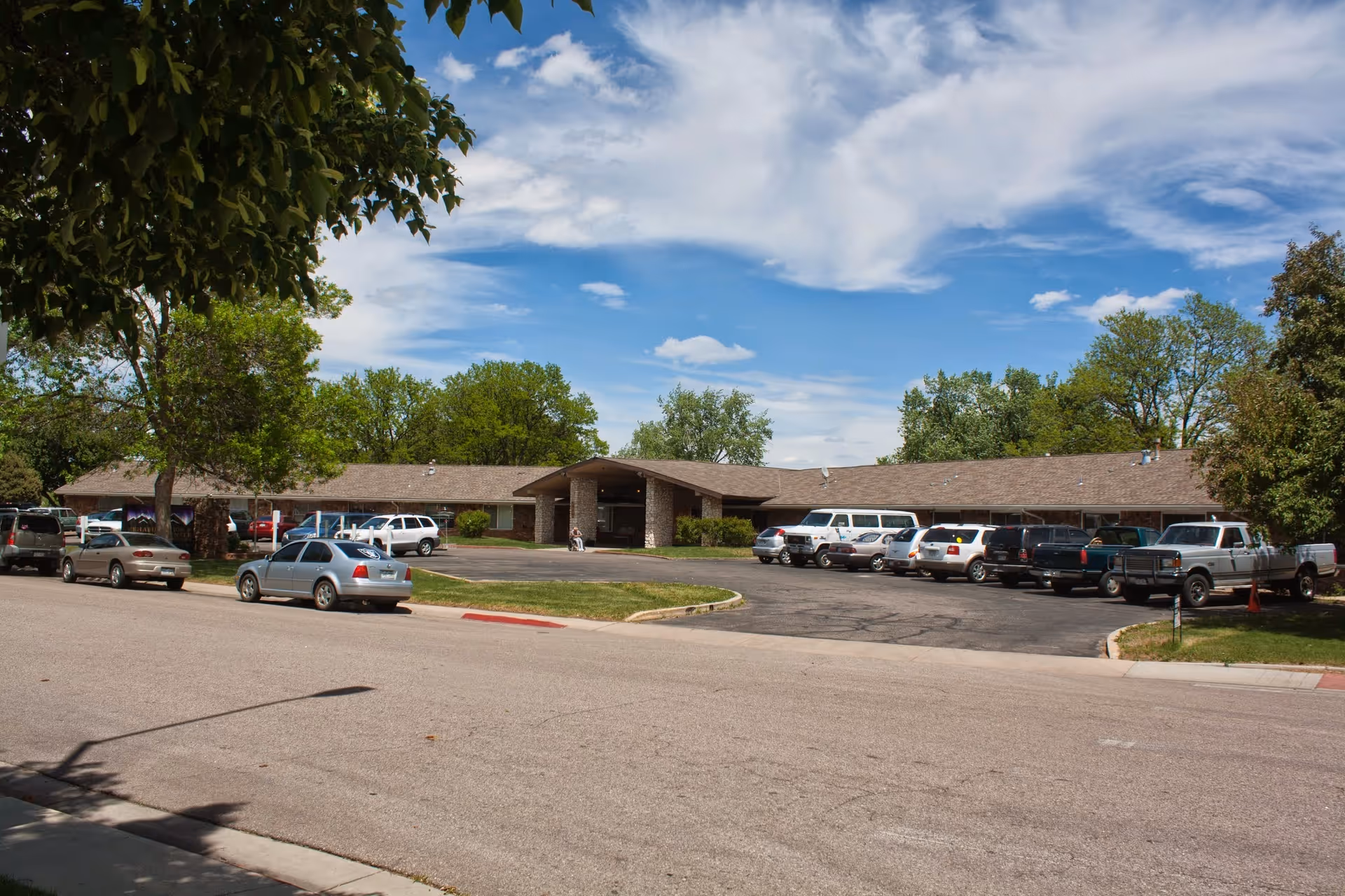 Exterior view of a single-story building with a sloped roof, surrounded by trees and a parking lot filled with cars under a partly cloudy blue sky.