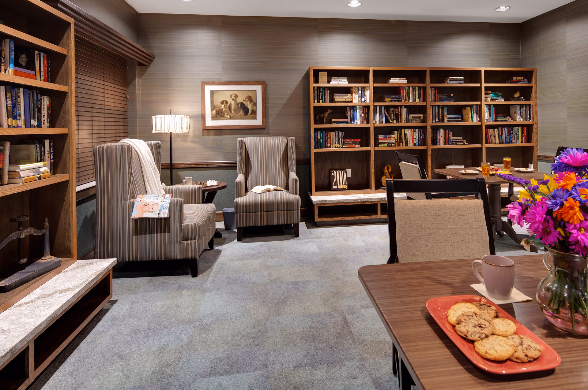 A cozy reading and sitting area in a senior living facility with two striped armchairs, a floor lamp, and wooden bookshelves filled with books. A table in the foreground has a vase of colorful flowers, a plate of cookies, and a coffee cup. The walls are decorated with a framed picture of dogs.