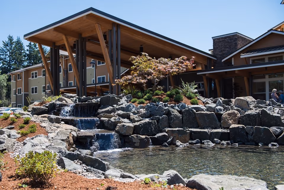Outdoor view of The Springs at Greer Gardens featuring a landscaped water feature with cascading waterfalls over rocks, surrounded by plants and mulch. In the background, there is a modern building with wooden beams and large windows under a clear blue sky.