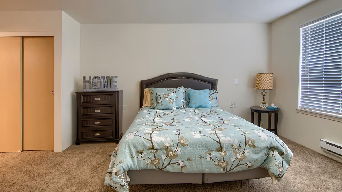 A neatly made bed with floral bedding is centered in a simple bedroom with a dresser, nightstand and window blinds.
