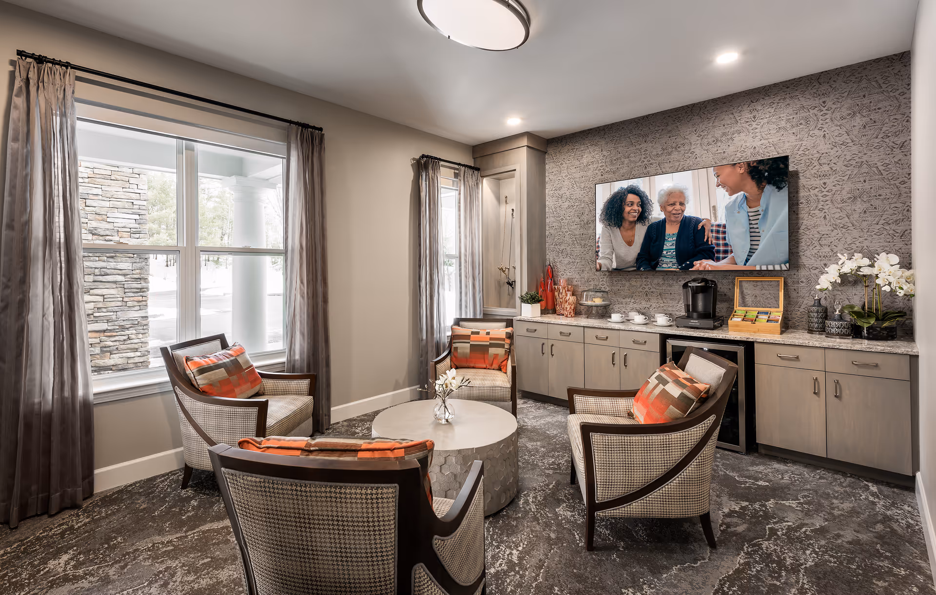 A cozy sitting area with four cushioned armchairs arranged around a round coffee table. The chairs have patterned upholstery and orange accent pillows. Behind the chairs is a long cabinet with a coffee maker, cups, and decorative items. A large flat-screen TV mounted on the wall displays an image of three women smiling and talking. The room has large windows with sheer curtains, allowing natural light to enter.