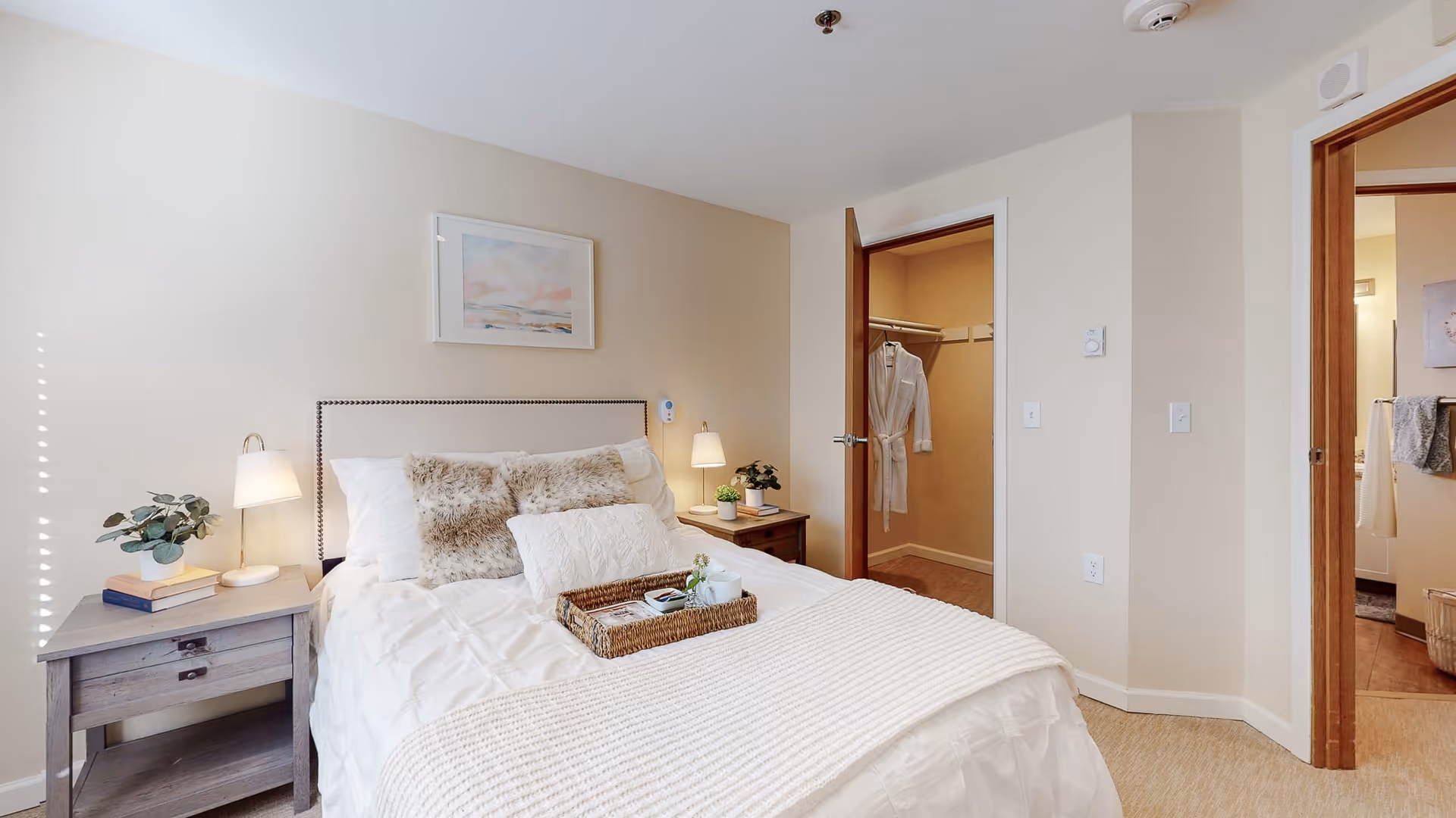 A cozy bedroom with a neatly made bed featuring white bedding and decorative pillows. There are two bedside tables with lamps, books, and plants. A framed artwork hangs above the bed. The room has beige walls and carpet flooring. An open door reveals a walk-in closet with a hanging robe, and another door leads to a bathroom.
