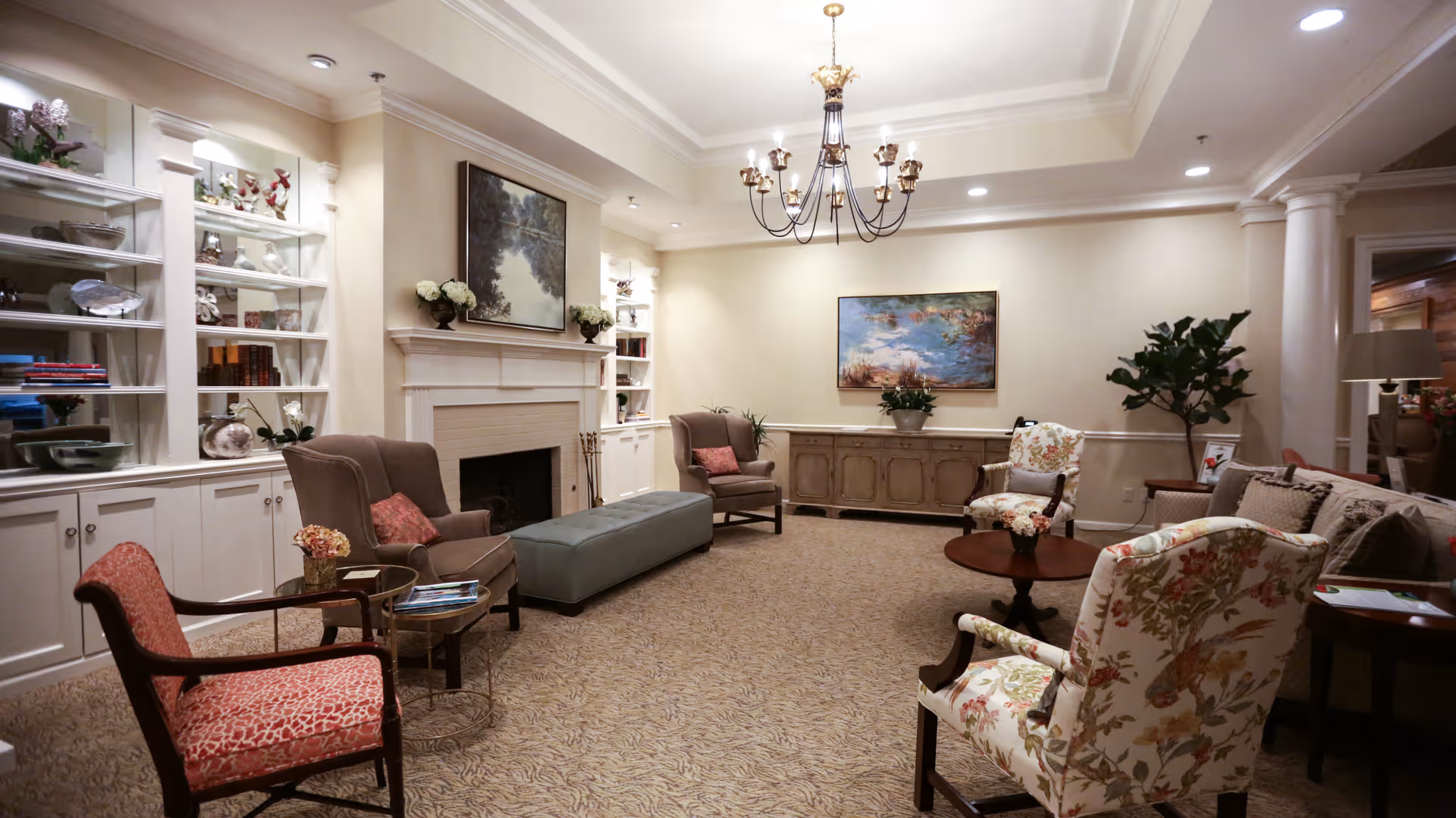 A cozy and elegant living room with beige walls and carpeted floor. The room features a white fireplace with a painting above it, built-in white shelves filled with decorative items and books, and a chandelier hanging from the ceiling. There are several upholstered chairs with floral and patterned fabric, a long cushioned bench, a round wooden coffee table with a flower arrangement, and a side table with a lamp. A large plant and another painting adorn the far wall.