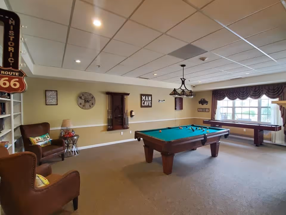 A recreational room with a pool table and a shuffleboard table. The room has beige walls with white trim, a ceiling with recessed lighting, and a large window with curtains letting in natural light. There are two brown leather armchairs with colorful pillows, a small side table with a lamp, and wall decorations including a clock, a 'Man Cave' sign, and Route 66 themed signs.