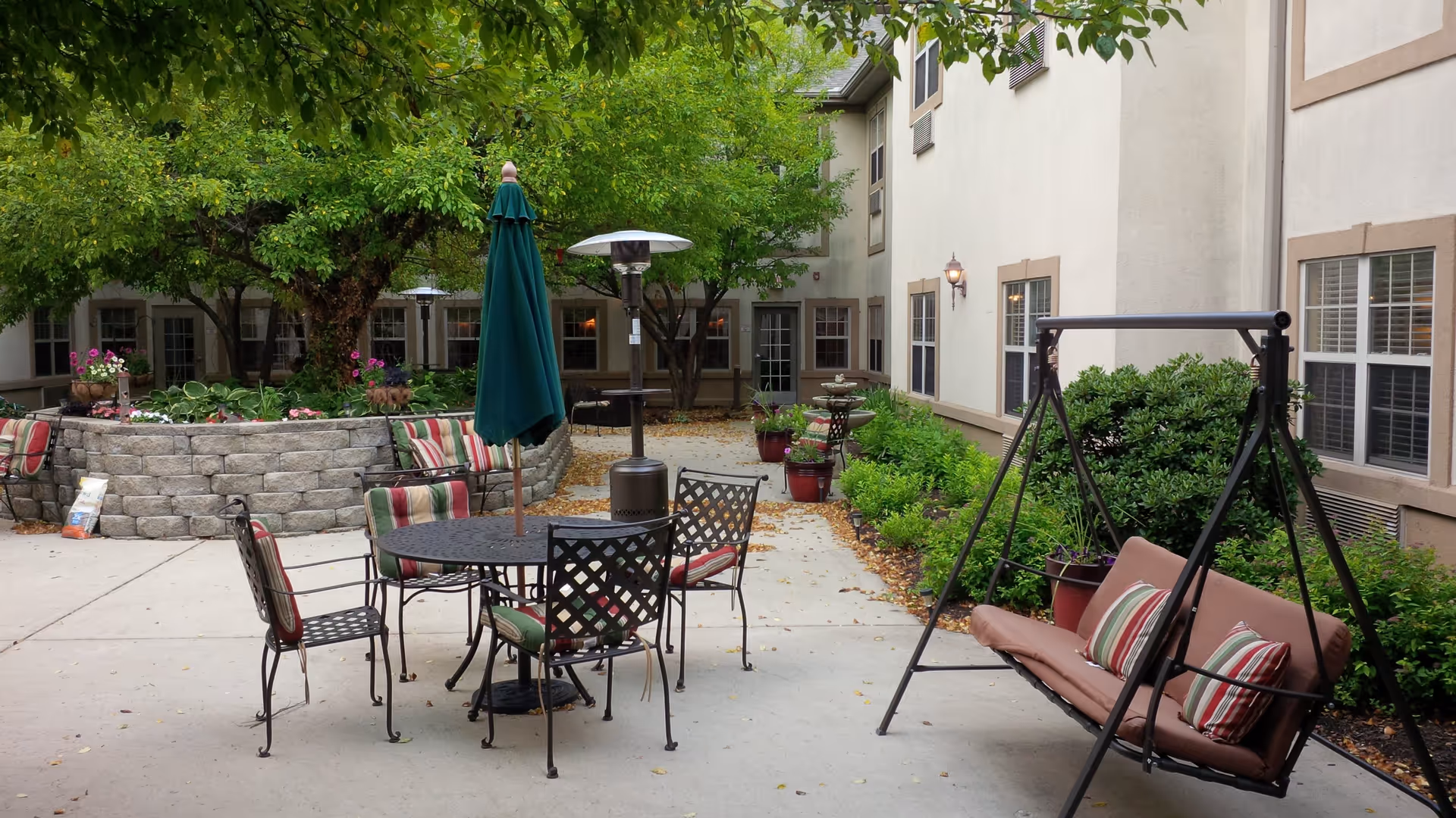 Outdoor patio area at Spring Mill Health Campus with a round metal table and four chairs with striped cushions, a closed green umbrella, a patio heater, a cushioned swing with striped pillows, potted plants, and trees surrounding the space.