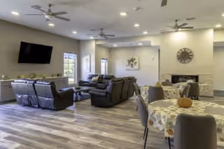 A spacious senior living common area with multiple seating arrangements including leather sofas and armchairs around a coffee table, a wall-mounted flat-screen TV, a fireplace with a large clock above it, and round dining tables covered with floral tablecloths and decorated with small pumpkins. The room has large windows, ceiling fans, and recessed lighting.