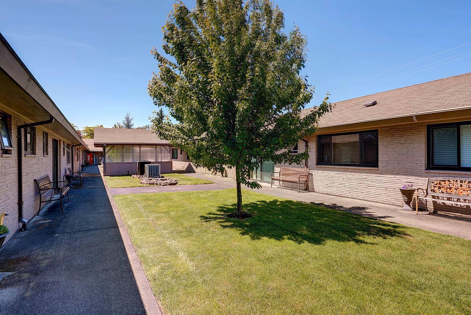 Outdoor courtyard area of Gateway Care & Retirement Center featuring a green lawn with a large tree in the center, surrounded by single-story buildings with beige brick walls and windows. Several benches are placed along the walkways under a clear blue sky.