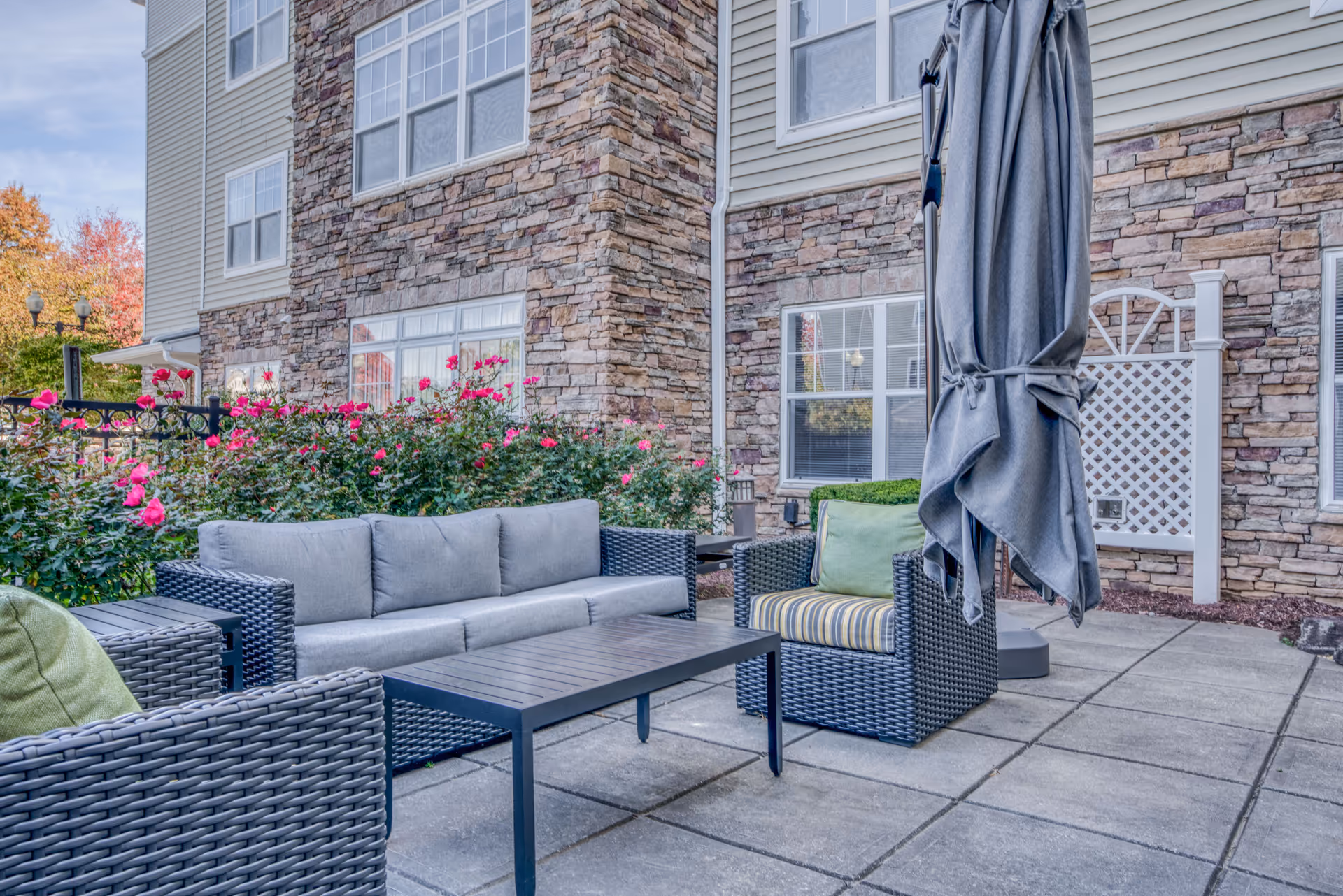Outdoor patio with wicker seating, a coffee table, closed umbrella, and flowering shrubs in front of a stone-faced senior living building.