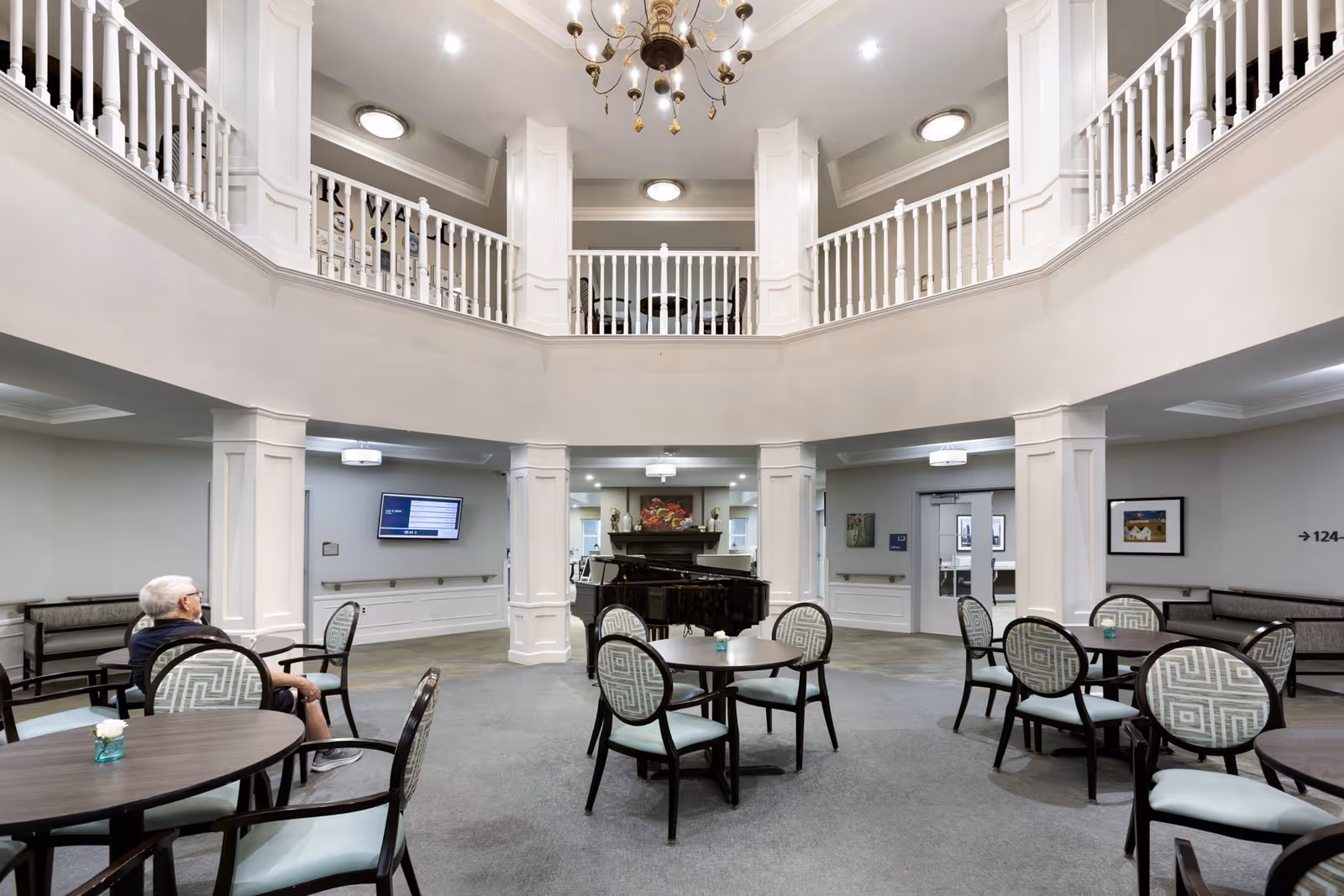 A spacious, well-lit common area in a senior living facility with round tables and patterned chairs arranged around the room. A grand piano is positioned near the center back wall, which features a fireplace and decorative artwork above it. The room has a two-story design with white railings on the upper level and a chandelier hanging from the ceiling. An elderly man is seated at one of the tables on the left side of the image.