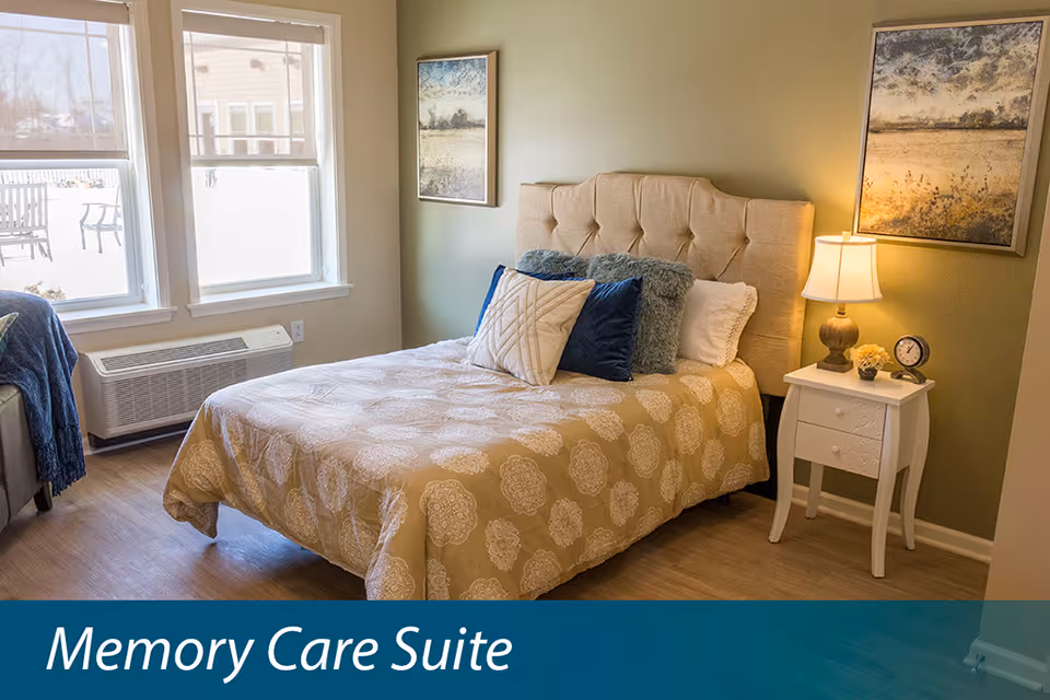 A cozy furnished bedroom with a bed, pillows, nightstand and lamp in a memory care suite.