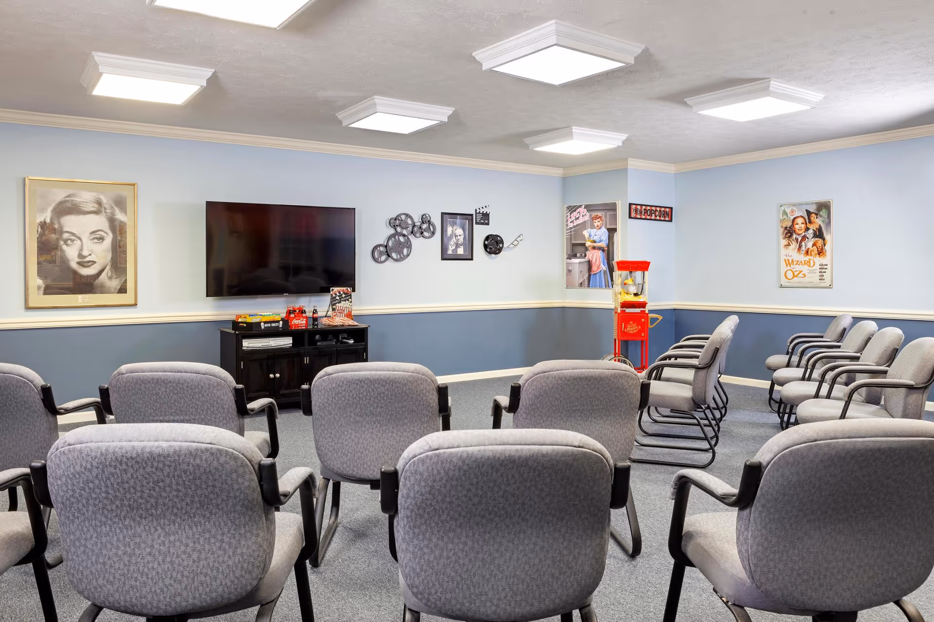 A small movie theater room with rows of gray cushioned chairs facing a flat-screen TV mounted on a light blue wall. The room features movie-themed decorations including framed vintage movie posters, film reels, and a red popcorn machine in the corner.