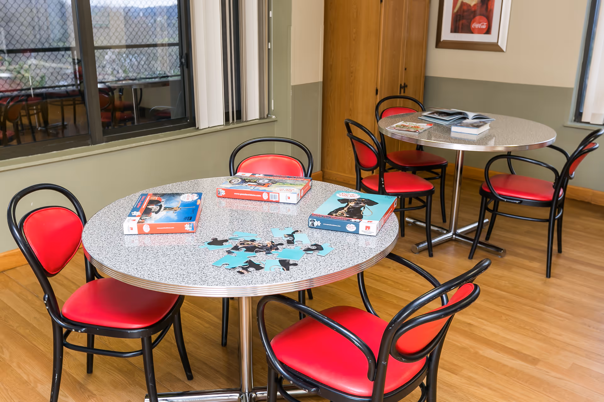 A room with two round tables surrounded by black chairs with red cushions. On one table, there are puzzle pieces and puzzle boxes, while the other table has some books and magazines. The room has wooden flooring, light green and beige walls, and windows with vertical blinds.