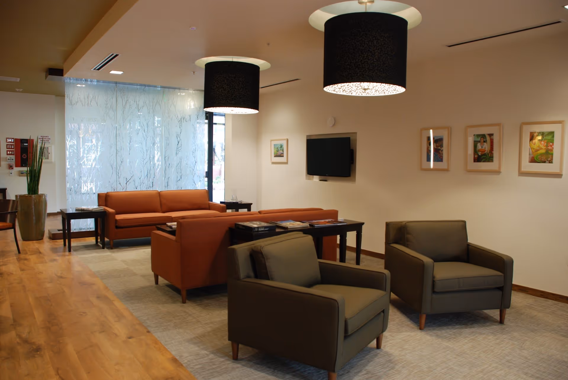 A modern living room area with two dark armchairs in the foreground and two orange sofas in the background. The room has wooden flooring, a beige carpet, and two large black pendant lights hanging from the ceiling. A flat-screen TV is mounted on the wall, which is decorated with four framed pictures. There is a large plant in a pot near a glass wall with frosted tree patterns.