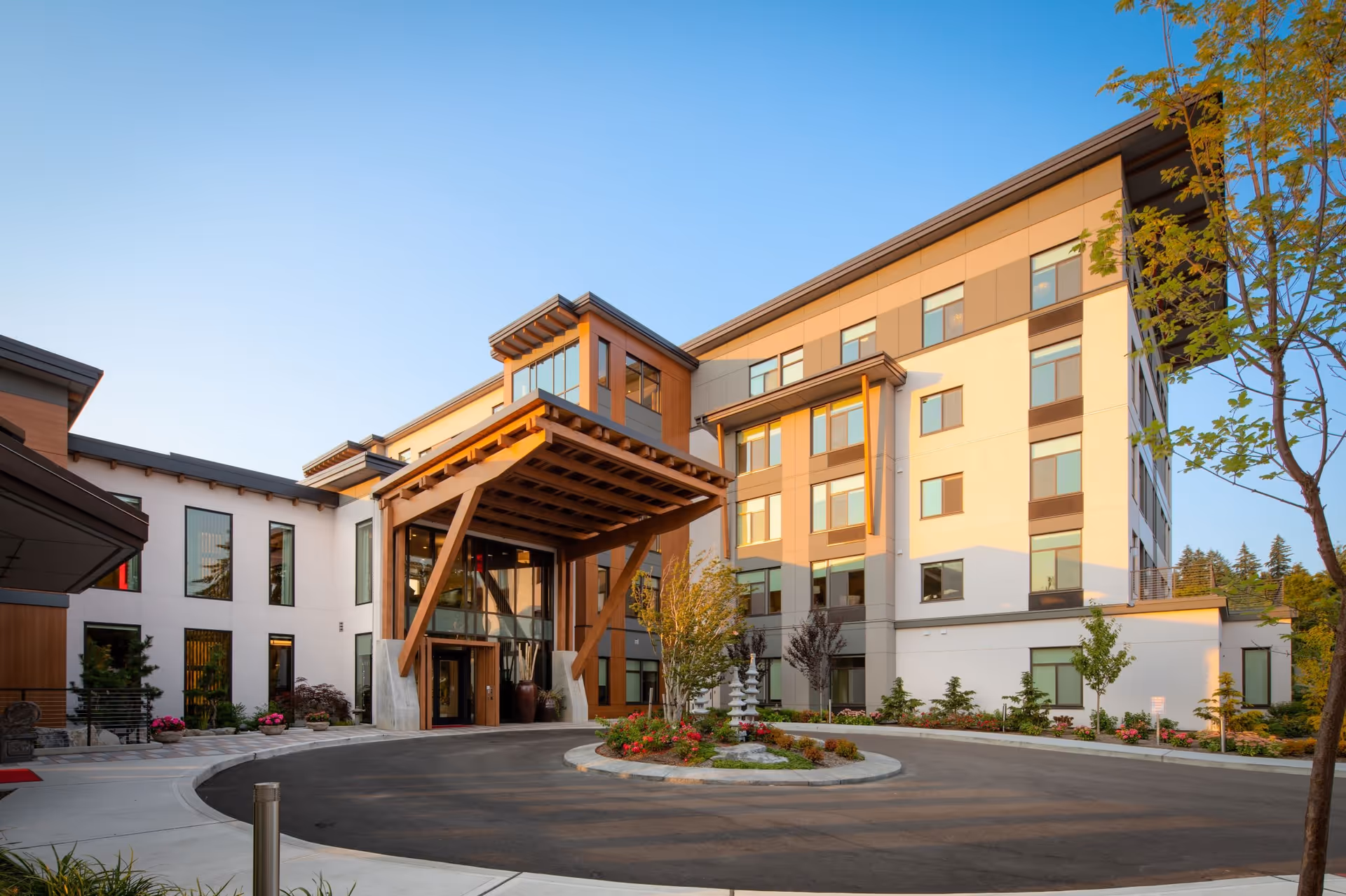 Exterior view of a modern multi-story senior living facility building with large windows, a covered entrance supported by wooden beams, landscaped garden beds, and a circular driveway under a clear blue sky.
