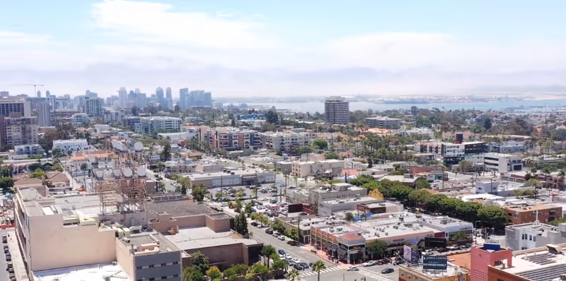 Aerial view of a cityscape showing numerous buildings, streets, and greenery with a body of water and distant mountains in the background under a partly cloudy sky.