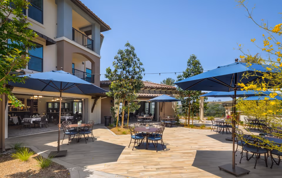 Outdoor patio area at Santianna Oakmont Signature Senior Living with multiple round tables and chairs under large blue umbrellas, surrounded by trees and plants, adjacent to a building with balconies and large windows.
