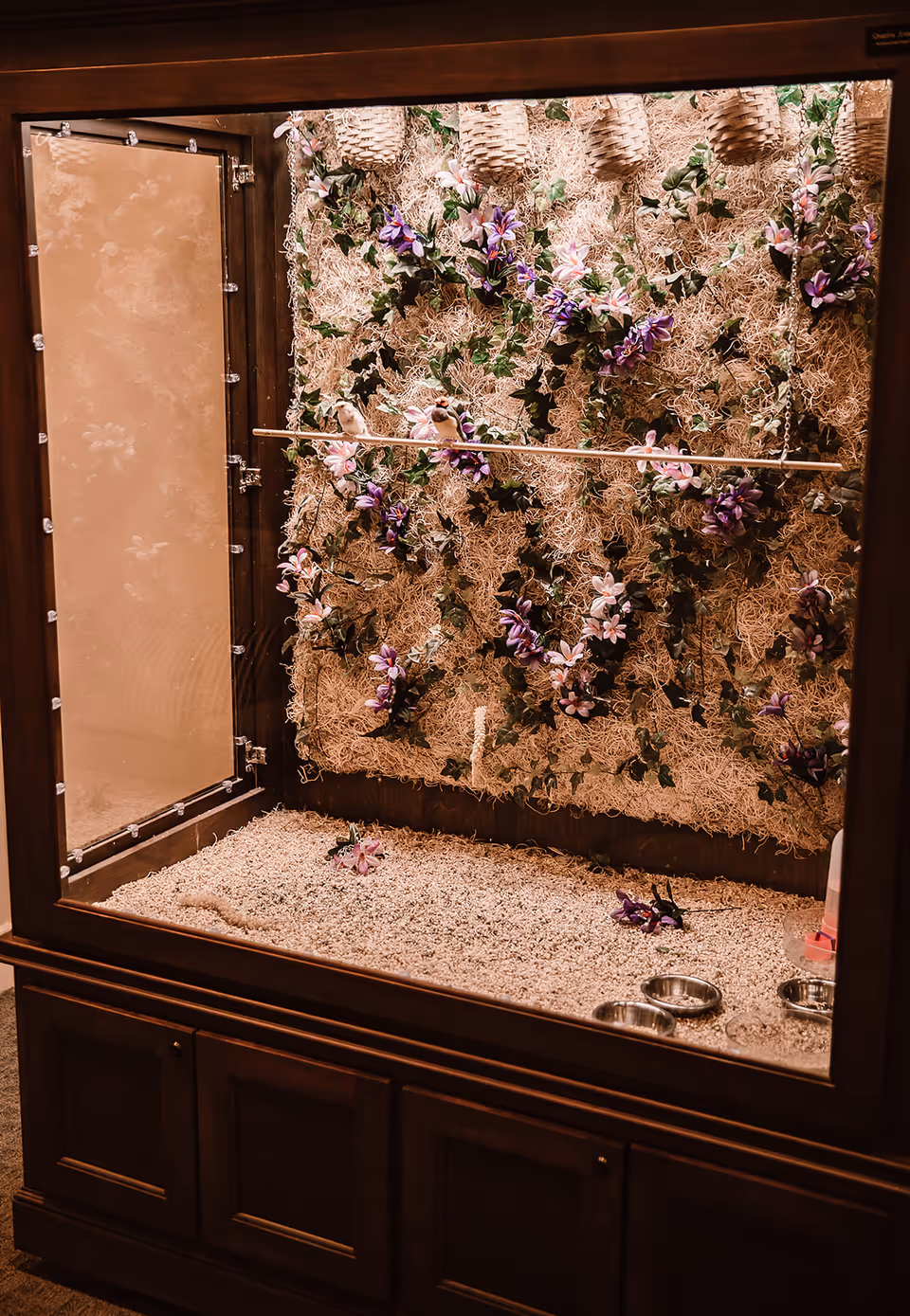 A wooden bird enclosure with a glass front, decorated inside with artificial flowers, vines, and hanging baskets. There are two small birds perched on a wooden rod inside the enclosure, which has a bedding of small pebbles or sand and metal food and water bowls on the floor.