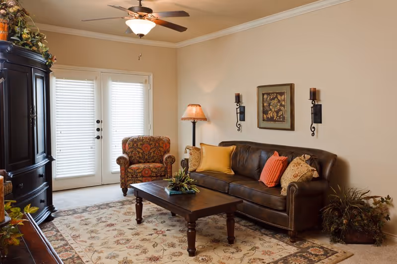 A cozy living room with a dark brown leather sofa adorned with colorful throw pillows, a patterned armchair, a wooden coffee table with a small plant centerpiece, a floor lamp, wall sconces, and a framed artwork on the beige wall. French doors with blinds allow natural light into the room, and a ceiling fan with a light fixture is mounted on the ceiling.