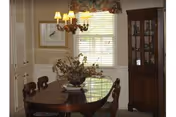 Dining room with a wooden table and chairs, a chandelier above a floral centerpiece, a window with blinds, and a china cabinet.