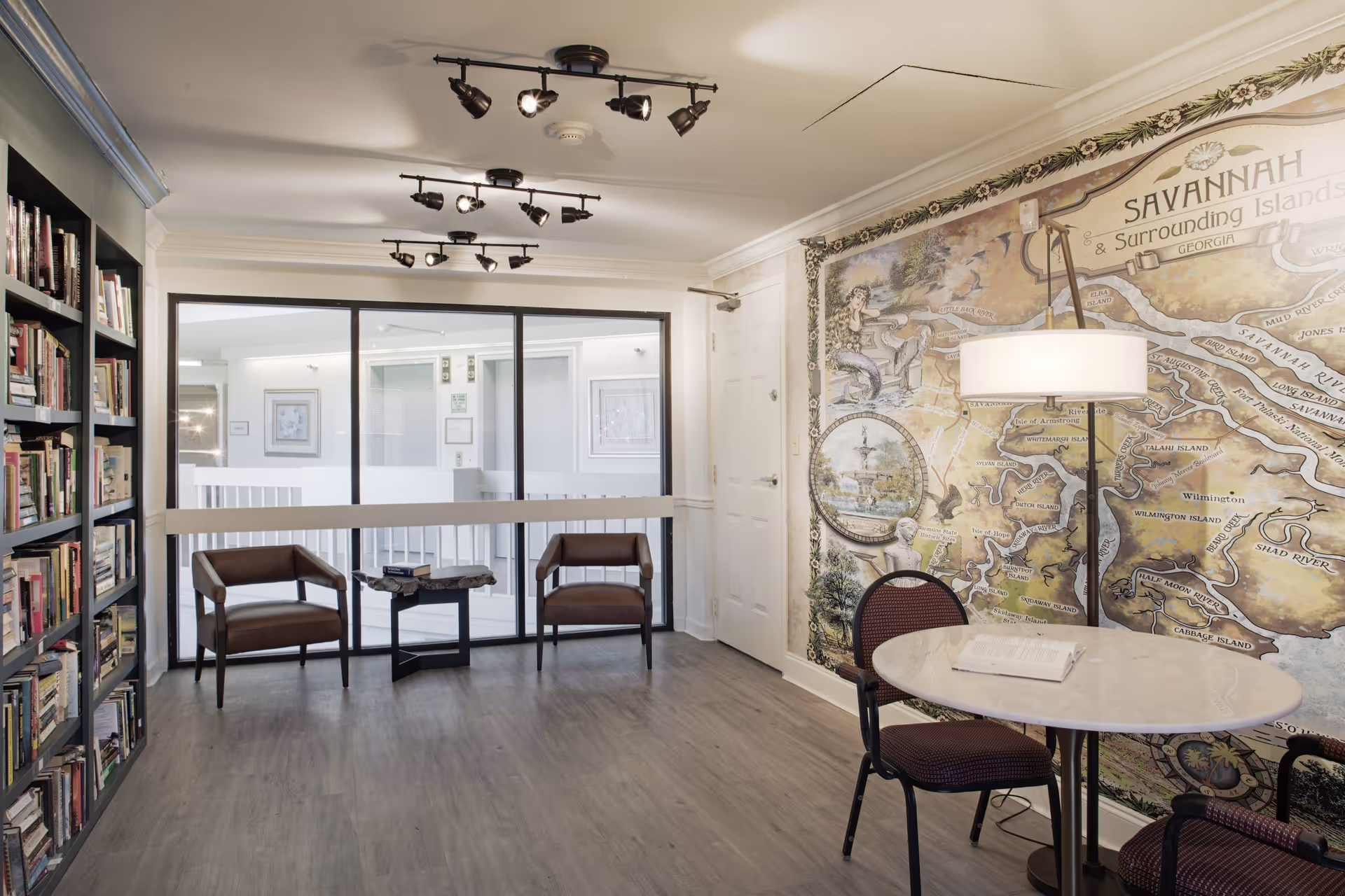 A cozy interior common area with a large bookshelf filled with books on the left, two brown leather chairs and a small table near a glass partition, and a round table with two chairs on the right. The wall behind the table features a large decorative map of Savannah and surrounding islands. The room has wood flooring and ceiling track lighting.