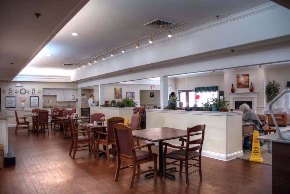 Interior view of a senior living facility dining area with several wooden tables and chairs arranged on a wooden floor. In the background, there is a kitchen area with white cabinets and a clock on the wall. To the right, there is a lounge area with a fireplace, plants, and seniors seated. The ceiling has recessed lighting and track lights.
