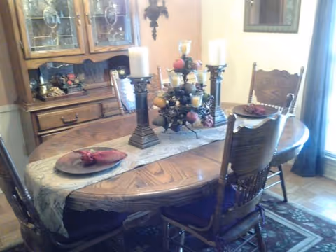 A wooden dining table set with four chairs in a dining room. The table has a decorative runner, two large candle holders with white candles, and a centerpiece with artificial fruit. In the background, there is a wooden china cabinet with glass doors displaying decorative items. A framed picture hangs on the wall, and a window with curtains is visible on the right side.