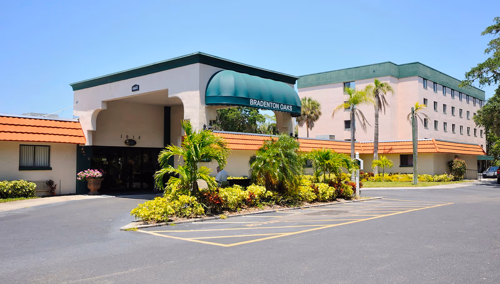 Exterior view of a senior living facility named Bradenton Oaks with a green awning over the entrance, surrounded by palm trees and landscaped bushes under a clear blue sky.
