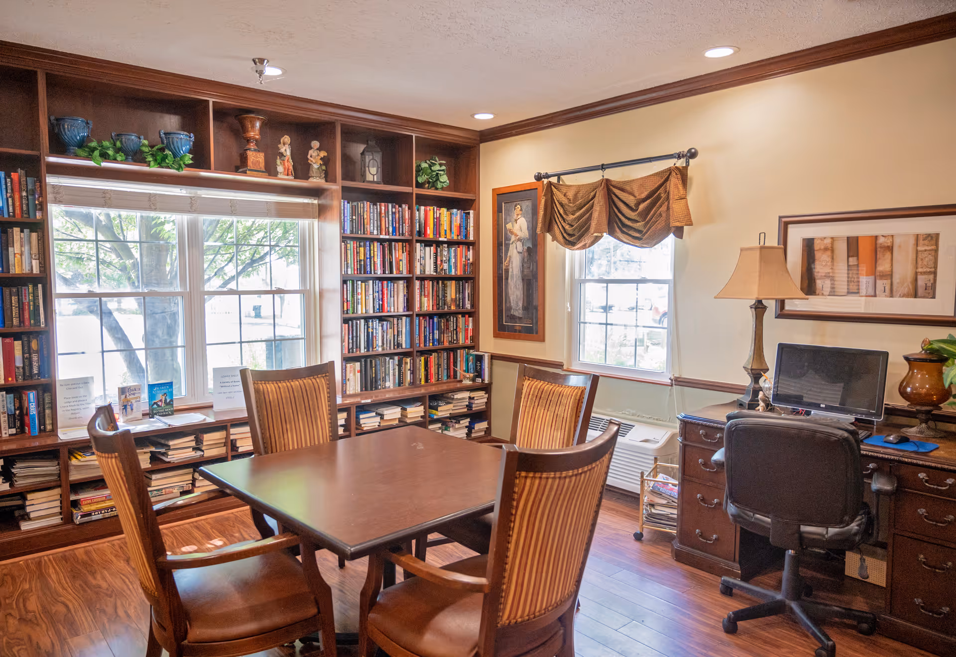 Sunlit library-style common room with a central table and four chairs, built-in bookshelves, windows, and a desk with a computer and lamp.