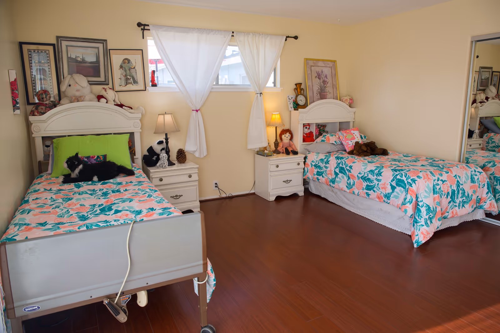 A cozy bedroom with two single beds featuring floral bedspreads in pink and green. Each bed has a white headboard and is accompanied by a white nightstand with a lamp and various stuffed animals and decorative items. The room has light yellow walls, a window with white curtains, and a wooden floor. A mirrored closet door is visible on the right side.