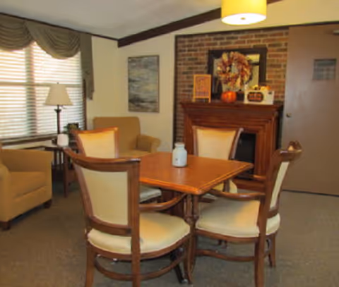 Cozy common room with a wooden table and four chairs, armchairs, a lamp, and a decorated fireplace.