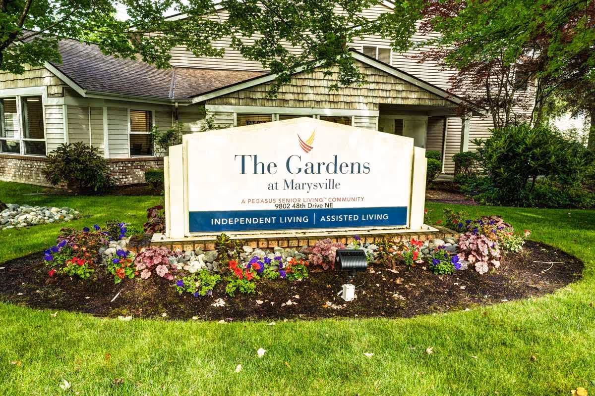 Outdoor view of the entrance sign for The Gardens at Marysville, a Pegasus Senior Living community offering independent and assisted living. The sign is surrounded by a flower bed with colorful flowers and greenery, with part of the building visible in the background.