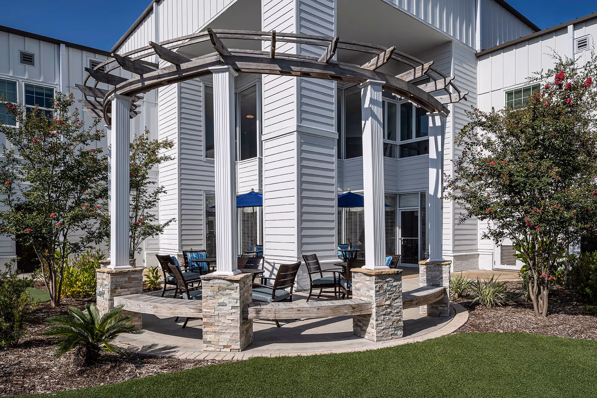 Outdoor seating area with a circular wooden pergola supported by white columns and stone bases, surrounded by chairs and tables with blue umbrellas. The area is landscaped with green grass, small plants, and flowering trees, adjacent to a white building with large windows.