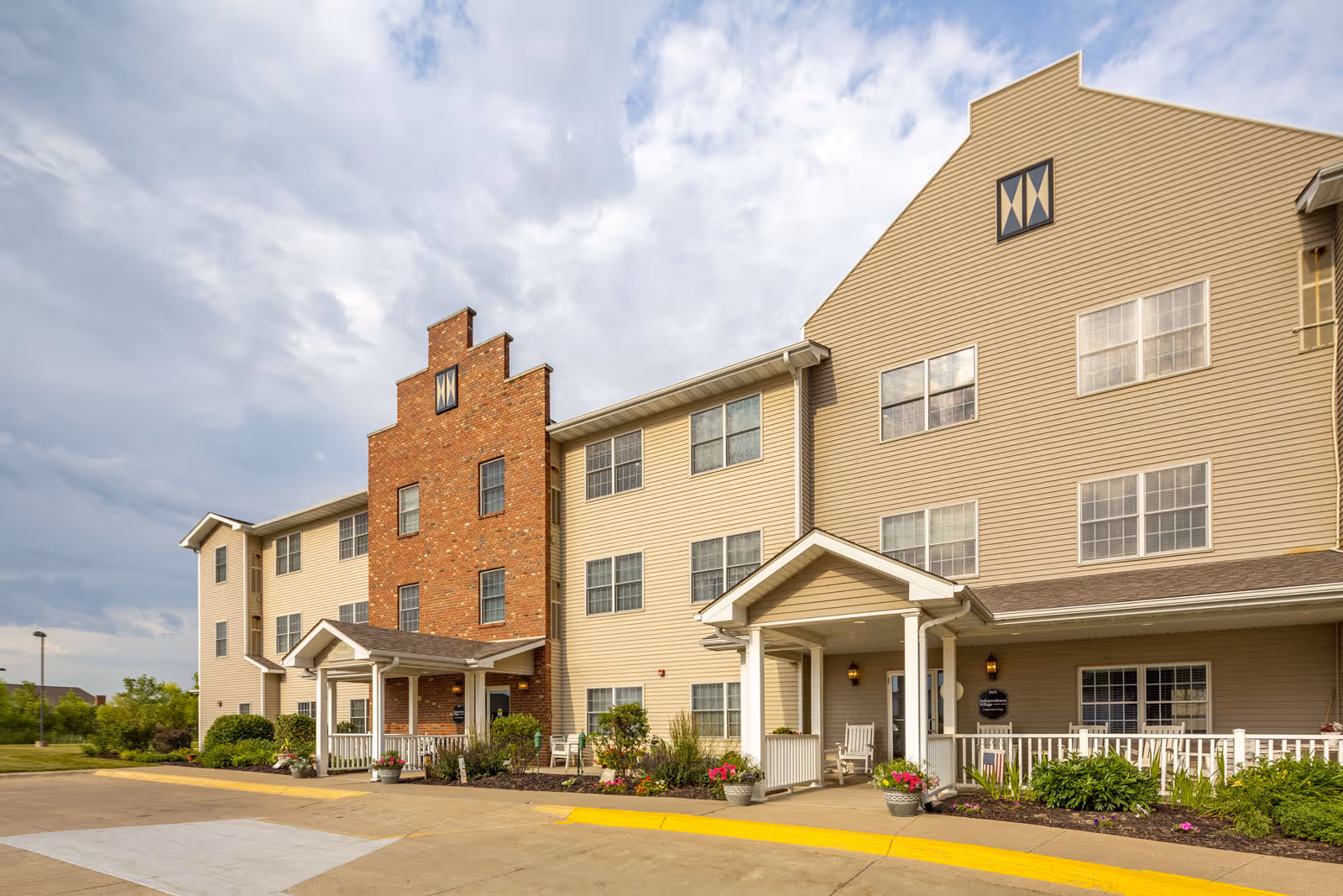 Exterior view of a multi-story senior living facility building with beige siding and a central section of red brick. The building has multiple windows and two covered entrances with white columns and railings. There are flower pots and landscaping along the front, and the sky is partly cloudy.