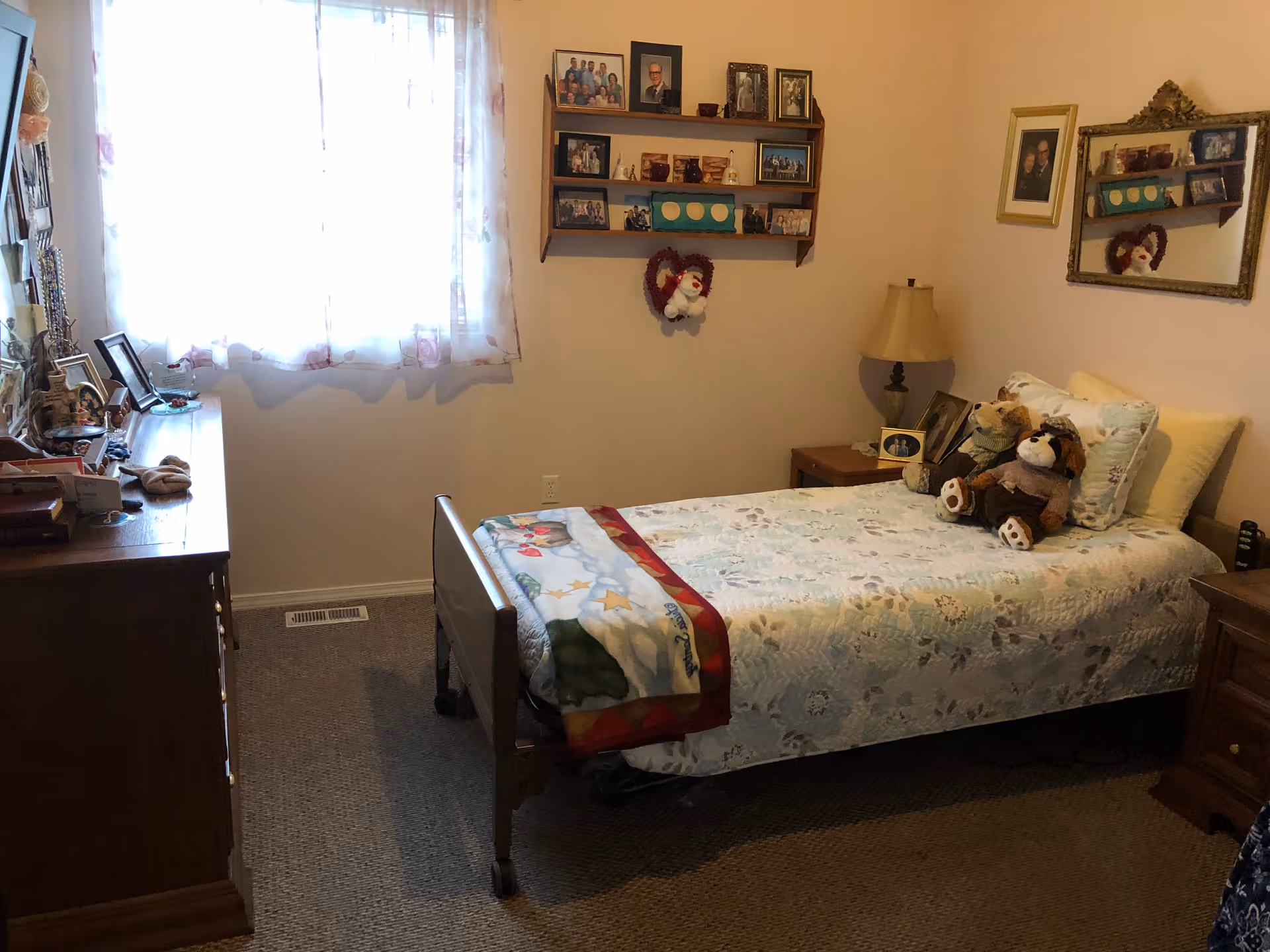 A cozy bedroom with a single bed covered in a floral quilt and a colorful blanket at the foot. The bed has several stuffed animals and pillows. There is a wooden dresser on the left side with various framed photos and items on top. A window with sheer curtains lets in natural light. On the wall above the bed, there are shelves with framed photos and decorative items. A bedside table with a lamp and more framed photos is next to the bed. A mirror hangs on the wall reflecting part of the room.
