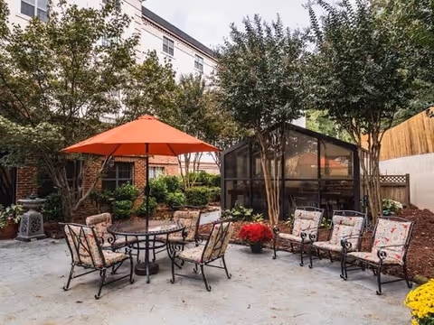 Outdoor patio area with a round table and six cushioned chairs under an orange umbrella. Nearby are four additional cushioned chairs arranged around a small table. The area is surrounded by trees, plants, and a small greenhouse structure. A multi-story brick building is visible in the background.