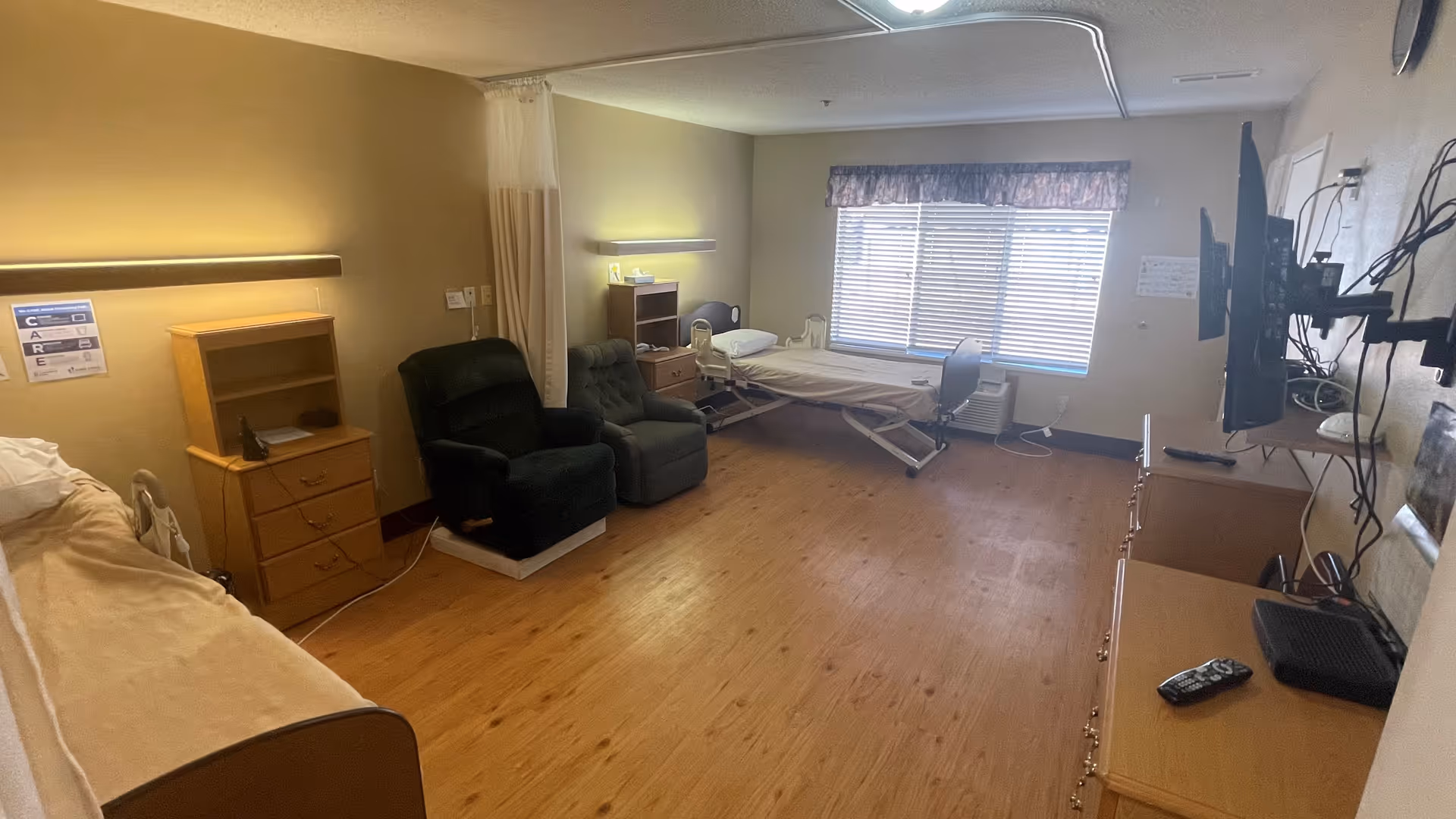 A spacious senior-care bedroom with two adjustable hospital beds, recliner chairs, wooden dressers, and a wall-mounted TV by a large window.
