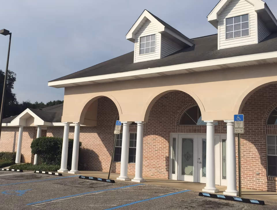 Front entrance of a brick building with arched portico, white columns, dormer windows, and marked handicap parking spaces.