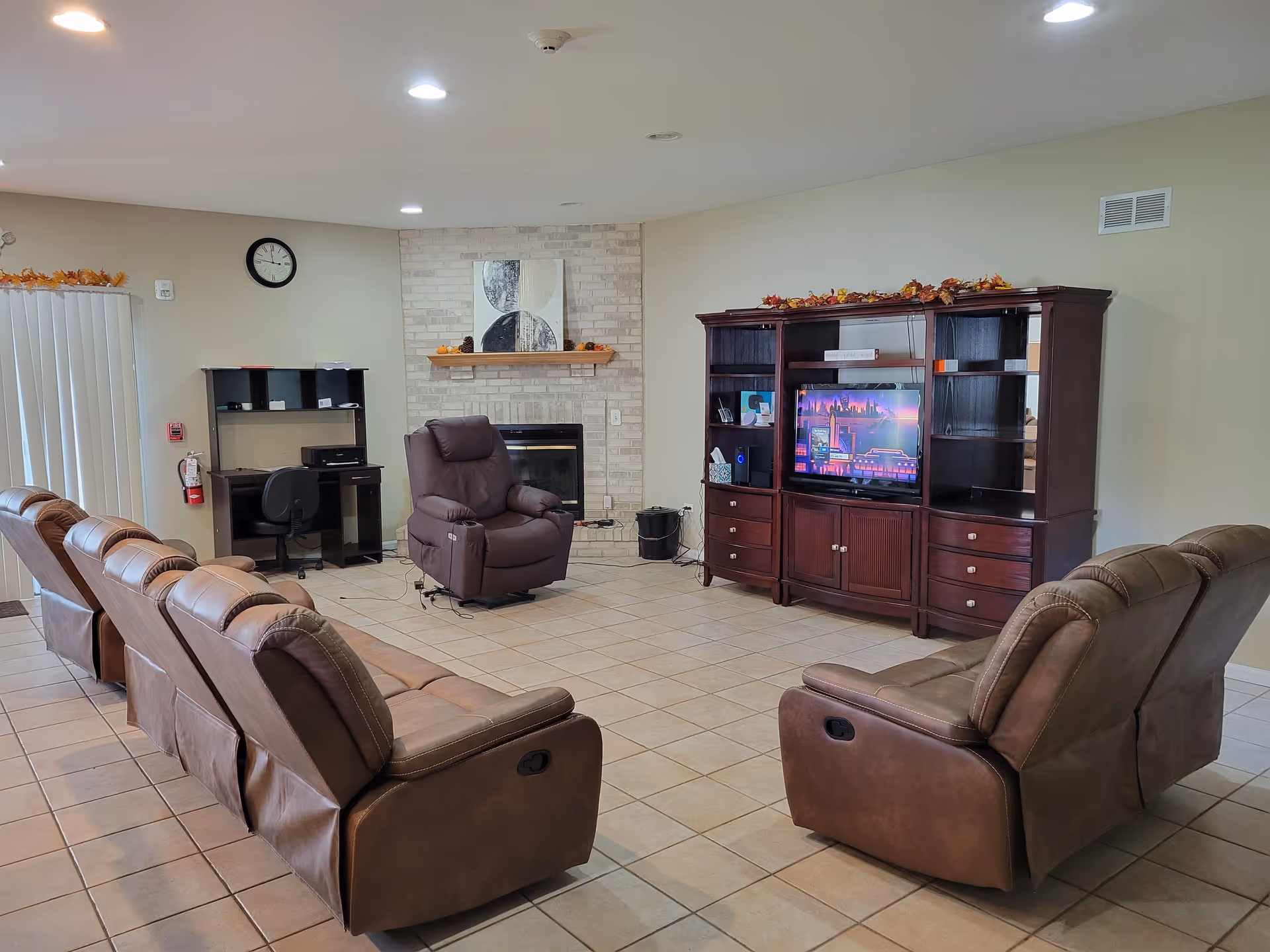 A senior living facility common area with several brown leather recliners arranged in a semi-circle facing a large wooden entertainment center with a TV. There is a fireplace with a decorative mantle and a piece of abstract art above it. A small desk with a chair and printer is positioned against the wall near vertical blinds covering a window or door. The room has tiled flooring and recessed ceiling lights.