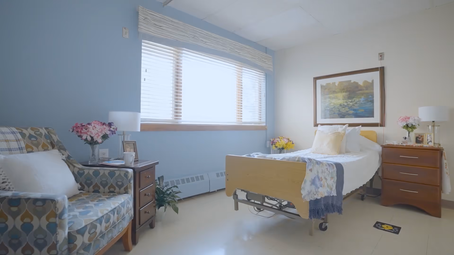 A bright and cozy bedroom in a senior living facility with a single bed covered in white bedding and a floral throw. There is a patterned armchair with a white pillow on the left, a wooden nightstand with a lamp, flowers, and picture frames on both sides of the bed. A large window with blinds lets in natural light, and a framed painting hangs on the wall above the bed.