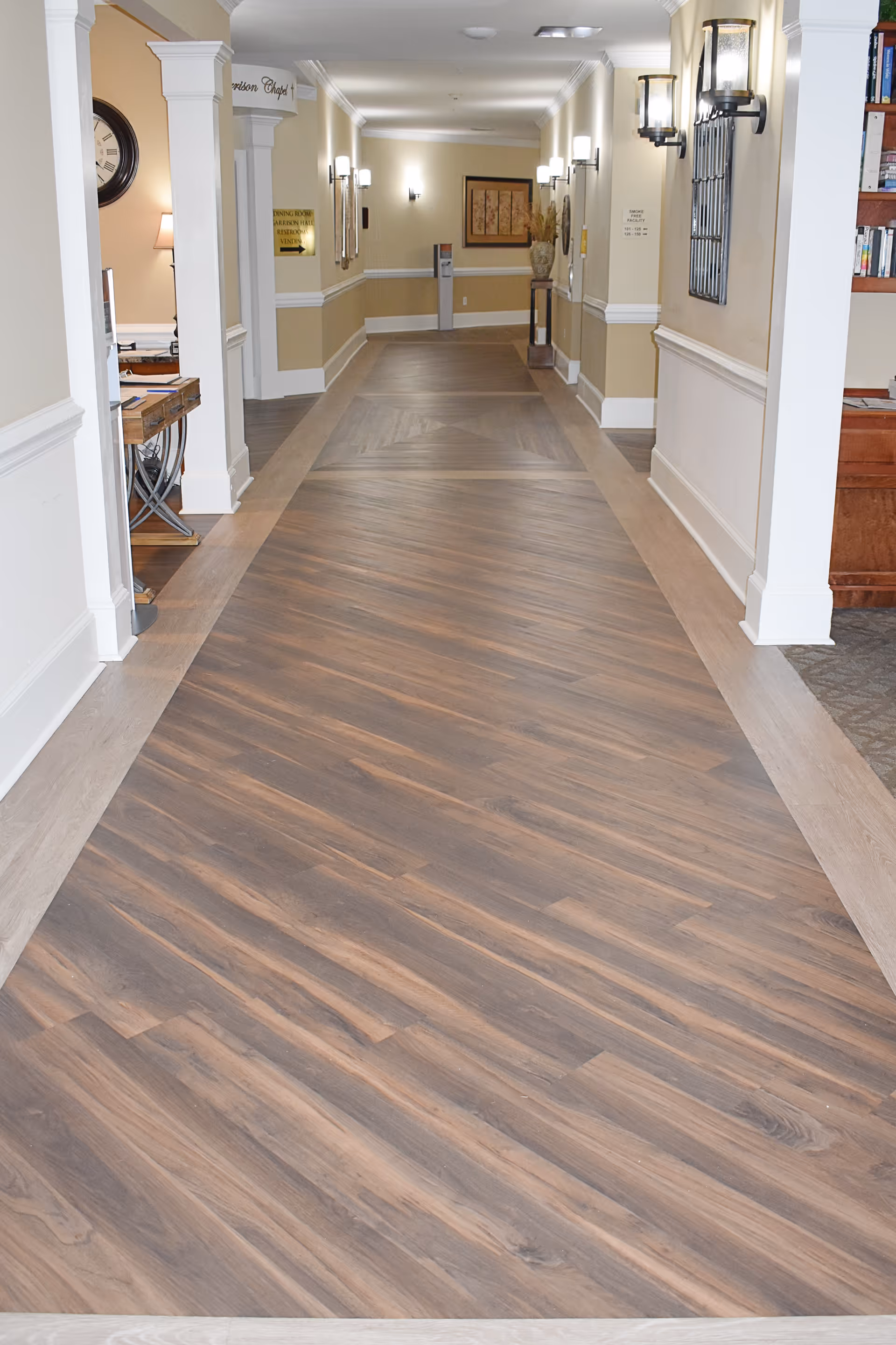 A well-lit hallway with wood-patterned flooring and beige walls with white trim. Wall sconces provide lighting along the corridor. On the left side, there is a glimpse of a room with a clock and a lamp on a desk. On the right side, there is a partial view of a bookshelf with books.