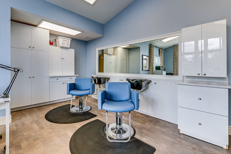 Interior view of a senior living facility hair salon with two blue salon chairs positioned in front of a large mirror. The room has light blue walls, white cabinetry, and two black hair washing sinks beneath the mirror. There are black floor mats under each chair and a laundry basket on top of the cabinets.
