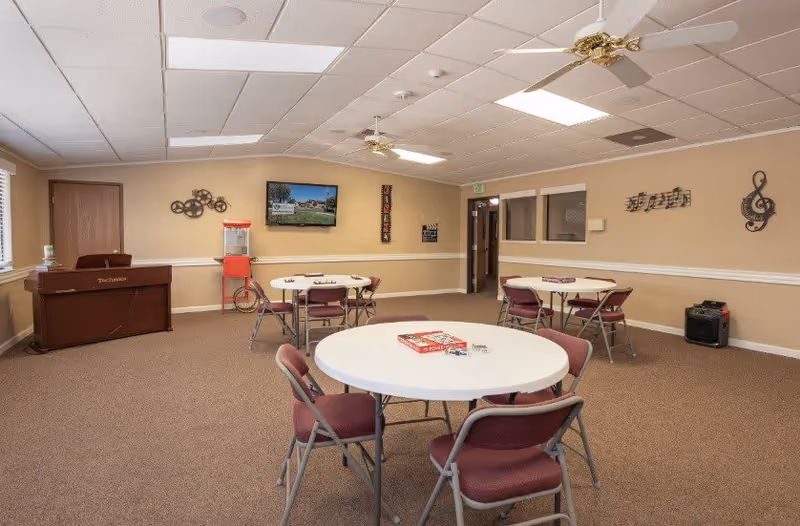 A senior living community activity room with round tables and chairs arranged for games. There are board games on the tables, a piano against the left wall, a popcorn machine, and wall decorations including musical notes and a treble clef. The room has beige walls, carpeted floor, and ceiling fans with lights.