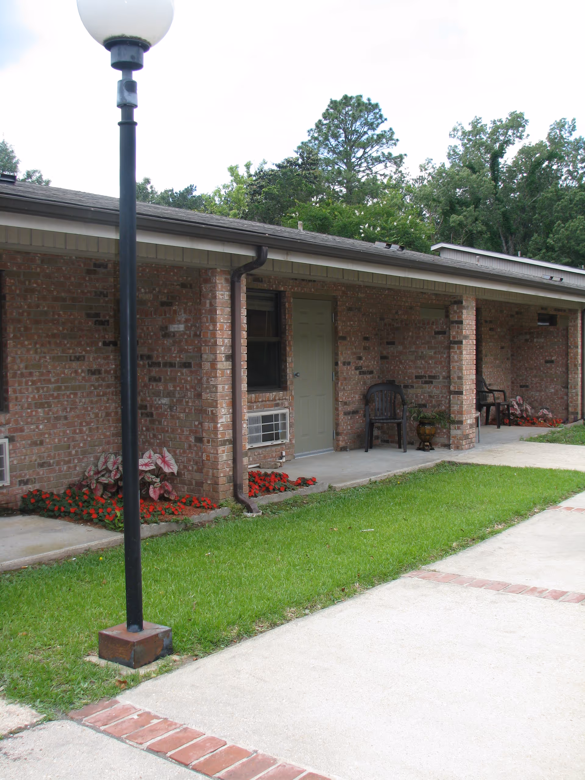 Exterior view of a single-story brick building with a covered walkway. There are two black chairs and potted plants near the doors, a lamp post in the foreground, and green grass with flower beds along the building. Trees are visible in the background.