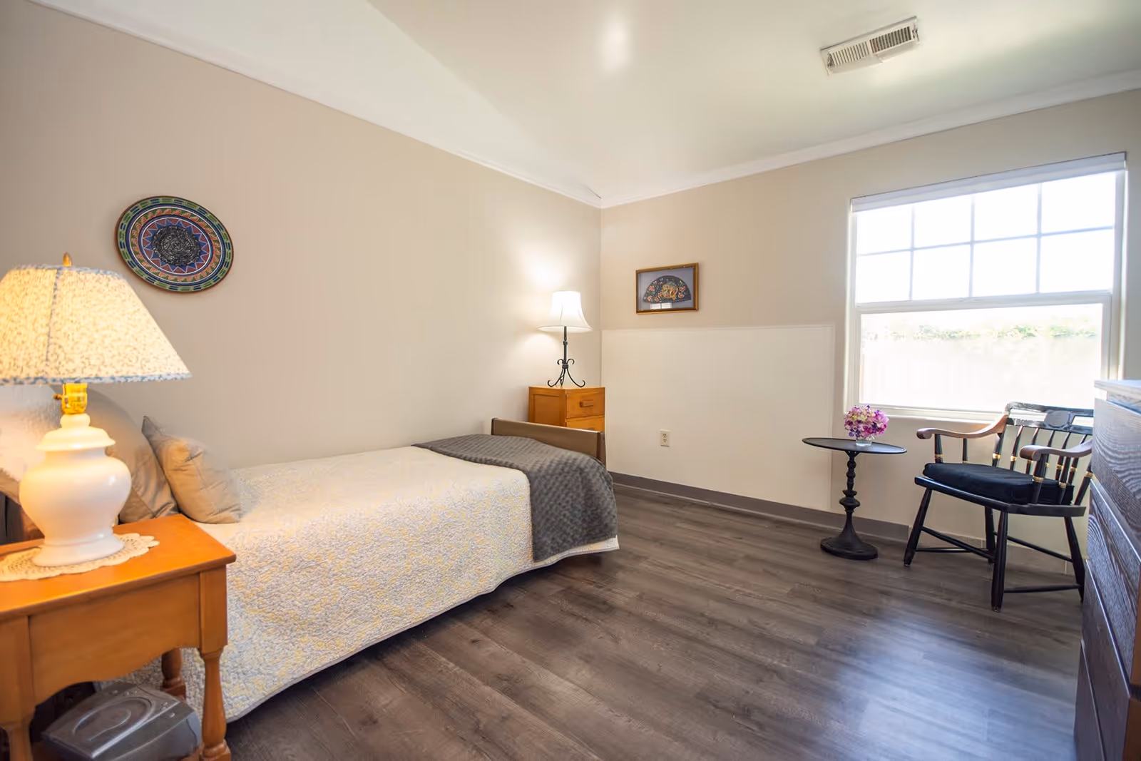 A simple, clean bedroom with a single bed covered with a light-colored quilt and a gray blanket. There is a wooden nightstand with a white lamp on it next to the bed. The room has light beige walls with a decorative plate and a framed picture hanging. A window with white blinds lets in natural light. A small round black table with a vase of purple flowers and a black wooden chair are near the window. The floor is dark wood laminate.