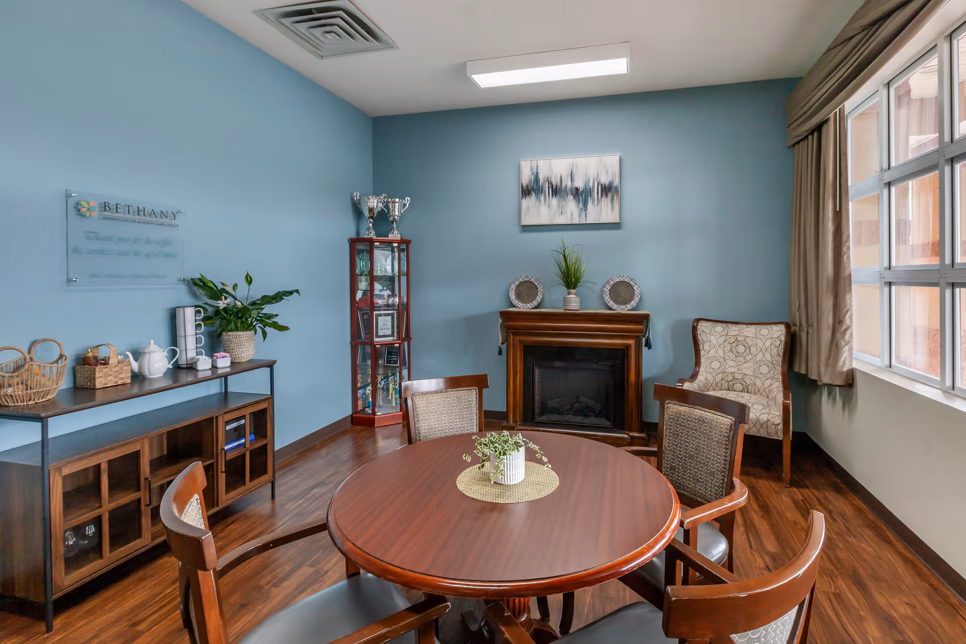 A cozy room with a round wooden table surrounded by four chairs. The walls are painted light blue, and there is a wooden cabinet with glass doors on the left side holding various items including a teapot and cups. A tall glass display cabinet with trophies and awards stands in the corner. A wooden fireplace mantel is decorated with two plates and a plant, and above it hangs an abstract painting. A large window with beige curtains lets in natural light on the right side.