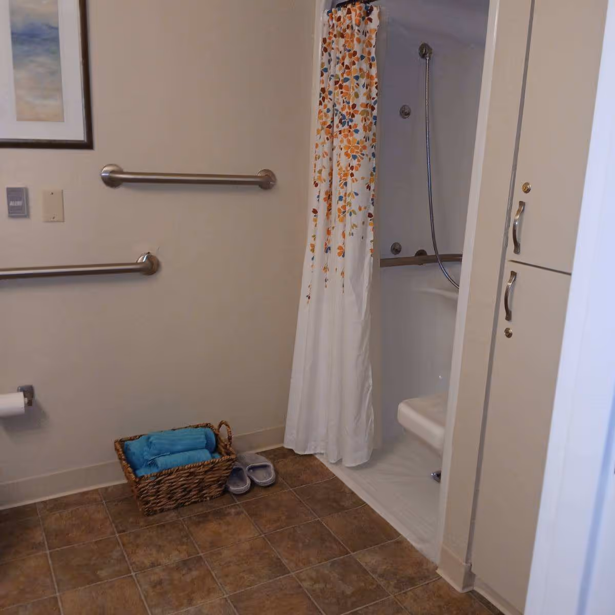 A bathroom with a tiled floor, a shower area with a floral-patterned shower curtain, grab bars on the wall, a wicker basket with blue towels, a pair of slippers on the floor, and a cabinet with silver handles.